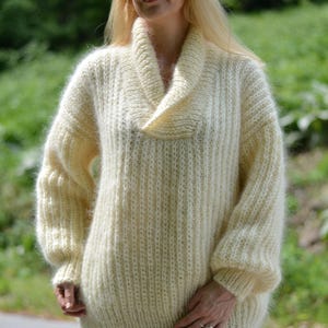 Op de afbeelding: Crèmekleurige, oversized trui met een brede, gedrapeerde col. De trui heeft een ribgebreide textuur en lange mouwen. Het model draagt een bril en staat buiten met een vage groene achtergrond.