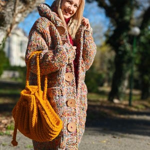 May include: A colourful, hooded, chunky knit cardigan with large wooden buttons and pockets. The cardigan is worn with a mustard-yellow drawstring bag. The garment appears to be handmade and suitable for cooler weather.