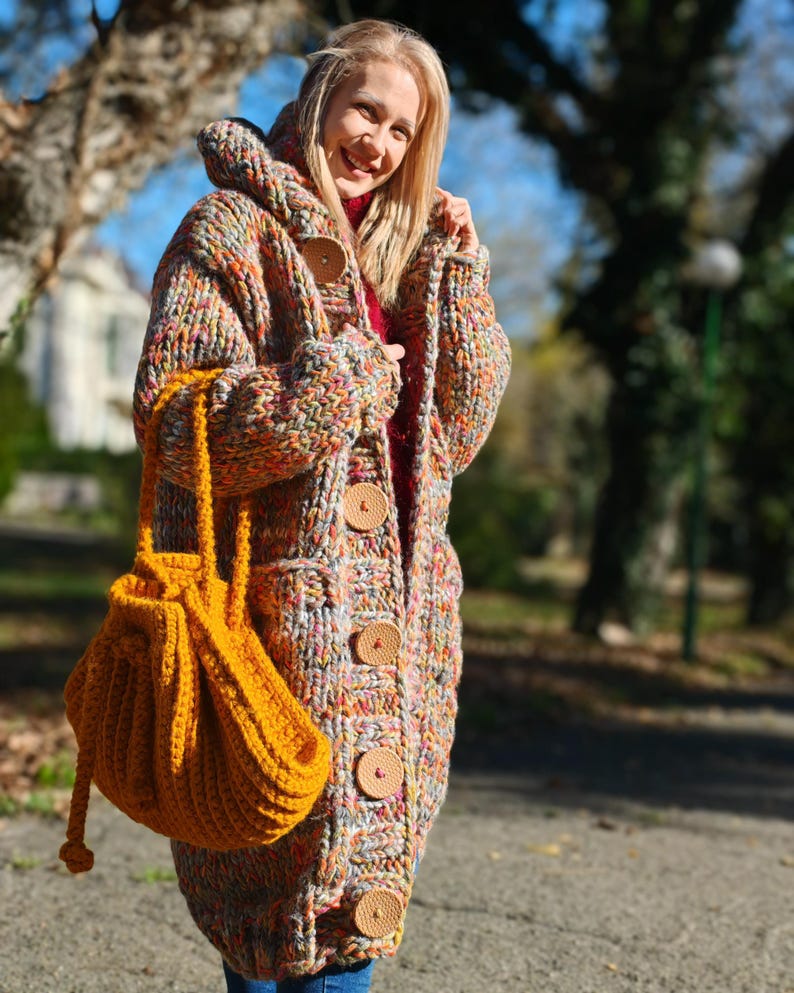 May include: A colourful, hooded, chunky knit cardigan with large wooden buttons and pockets. The cardigan is worn with a mustard-yellow drawstring bag. The garment appears to be handmade and suitable for cooler weather.