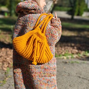 May include: A person wearing a long, hooded, multi-coloured knitted coat and carrying a bright yellow knitted tote bag. The coat is a mix of grey, orange, pink, and yellow yarns. The tote bag is a solid mustard yellow colour.