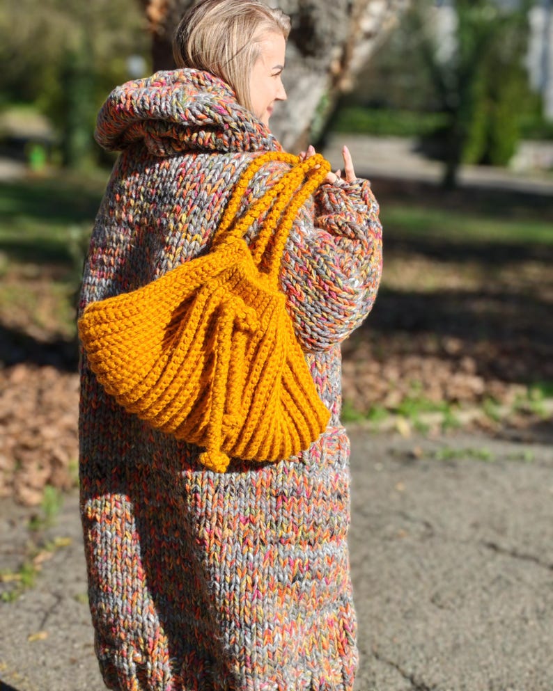 May include: A person wearing a long, hooded, multi-coloured knitted coat and carrying a bright yellow knitted tote bag. The coat is a mix of grey, orange, pink, and yellow yarns. The tote bag is a solid mustard yellow colour.