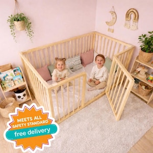 May include: A light-colored wooden playpen with vertical slats, featuring two children inside. The playpen has an open gate and is set on a white rug. The image includes a shelf with books and toys, and the text "MEETS ALL SAFETY STANDARD".