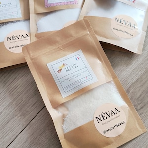Pode incluir: Vários pequenos sacos de papel castanho cheios de pó branco estão dispostos numa superfície de madeira. Cada saco tem uma etiqueta com texto e um autocolante "NÉVAA". Os sacos contêm pó perfumado para aspiradores.