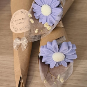 Può includere: Due bouquet di saponi a forma di margherita color lavanda con centri bianchi, avvolti in carta marrone e delicato tessuto bianco. Un bouquet ha un adesivo con "Bonne fête Maman" e un piccolo fiocco.