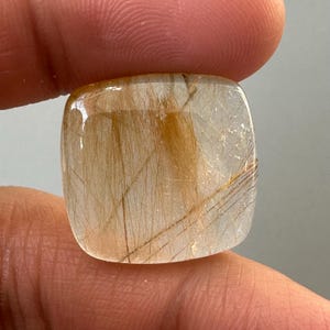 May include: A square-cut gemstone with a clear base and golden-brown rutile inclusions. The gemstone is held between fingers, showcasing its polished surface and the intricate patterns within. The stone's shape is a rounded square, and the inclusions create a striking visual effect.