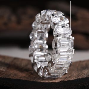 May include: A silver-toned eternity band featuring alternating oval and emerald-cut lab-grown stones. The ring displays a continuous pattern of clear, faceted gems set in a metal frame. Text on the image indicates Lab-Grown: 7.00 mm x 5.00 mm.