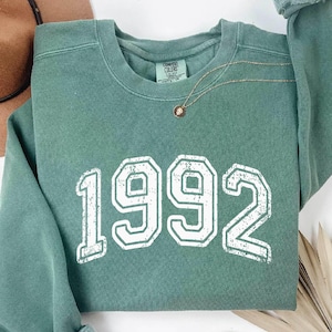 May include: A sage green sweatshirt with the year "1992" printed in white, with a distressed font. A gold necklace with a small pendant rests on the shirt. A brown hat and dried plant are in the background.