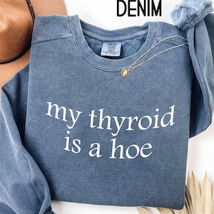 May include: Denim blue sweatshirt with the text "my thyroid is a hoe" in white. The sweatshirt has a crew neck and long sleeves. A gold necklace with a pendant is draped across the front.