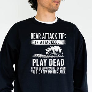 May include: Black sweatshirt with white text: "BEAR ATTACK TIP: IF ATTACKED, PLAY DEAD. IT WILL BE GOOD PRACTIC FOR WHEN YOU DIE A FEW MINUTES LATER." A bear illustration is also featured.