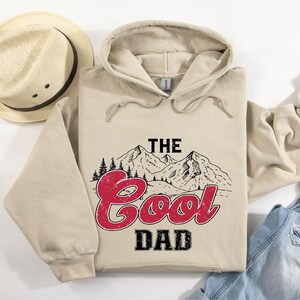May include: A beige hoodie with the text "THE Cool DAD" in red and black, featuring a mountain and tree graphic. A straw hat with a dark brown band is to the left of the hoodie. The hoodie is folded on top of a pair of blue jeans.