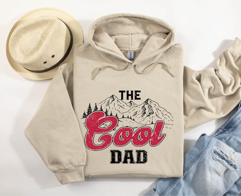May include: A beige hoodie with the text "THE Cool DAD" in red and black, featuring a mountain and tree graphic. A straw hat with a dark brown band is to the left of the hoodie. The hoodie is folded on top of a pair of blue jeans.