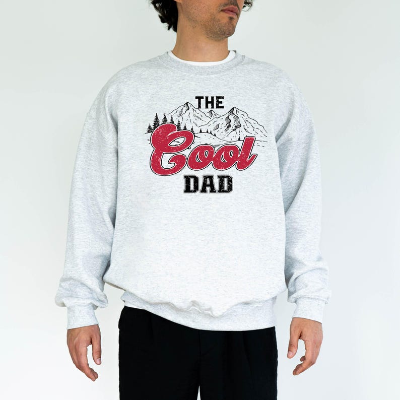 May include: A light gray sweatshirt with the text "THE Cool DAD" in a red, black, and white graphic. The graphic features a mountain range and trees. The sweatshirt has long sleeves and a crew neck.