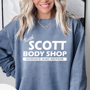 May include: A blue-grey sweatshirt with the text "Keith SCOTT BODY SHOP SERVICE AND REPAIR" in white. The sweatshirt has long sleeves and a crew neck.