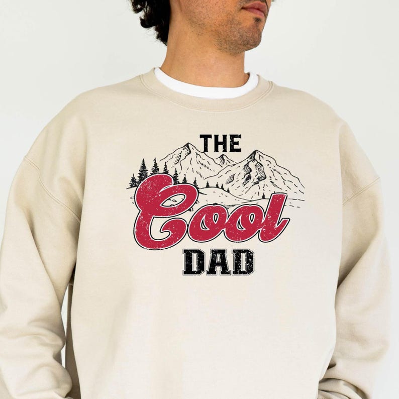 May include: A beige sweatshirt with a white crew neck collar. The front features a graphic of mountains and the text "THE Cool DAD" in red and black.