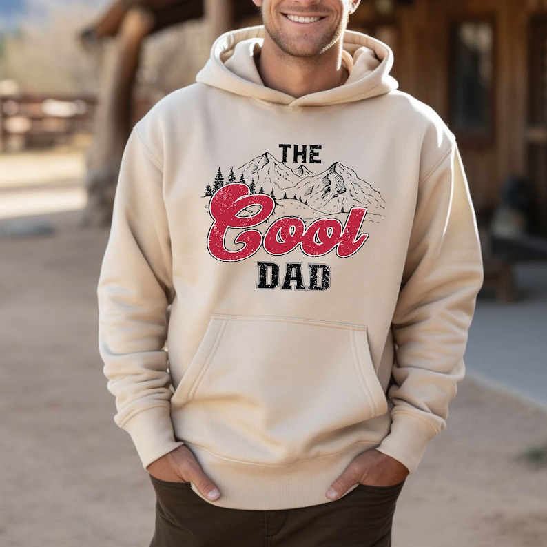 May include: A beige hoodie with a front pocket and a drawstring hood. The front features a graphic with the text "THE Cool DAD" in red and black, along with a mountain and tree design.