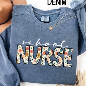 May include: A denim blue sweatshirt with the words "school NURSE" in a floral design. The sweatshirt has a crew neck and long sleeves. A gold necklace with a pendant is draped on the shirt.