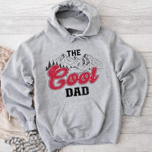 May include: A gray hooded sweatshirt with a front pocket and the words "THE Cool DAD" in a mountain-themed design. The text "Cool" is in red, and the word "DAD" is in black.