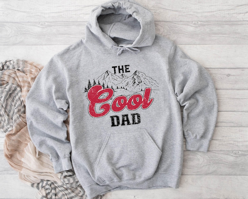 May include: A gray hooded sweatshirt with a front pocket and the words "THE Cool DAD" in a mountain-themed design. The text "Cool" is in red, and the word "DAD" is in black.