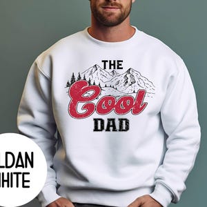 May include: White sweatshirt with the text "The Cool Dad" in a distressed red font, with a mountain and pine tree graphic. The sweatshirt has a crew neck and long sleeves. The brand name "Gildan White" is printed on a white circle.
