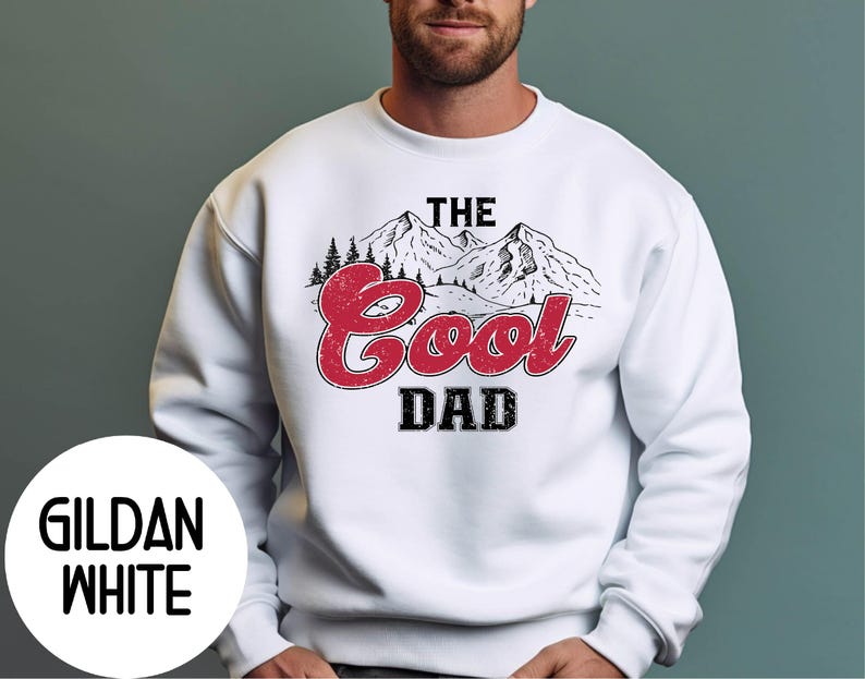 May include: White sweatshirt with the text "The Cool Dad" in a distressed red font, with a mountain and pine tree graphic. The sweatshirt has a crew neck and long sleeves. The brand name "Gildan White" is printed on a white circle.