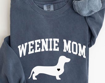 May include: A slate blue sweatshirt with the words "WEENIE MOM" in white block letters and a white dachshund silhouette with a black heart. The sweatshirt has a crew neck and long sleeves.