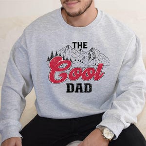 May include: A light gray sweatshirt with the text "THE Cool DAD" in a red and black design. The graphic includes a mountain range and trees. The sweatshirt is long-sleeved and made of a soft material.