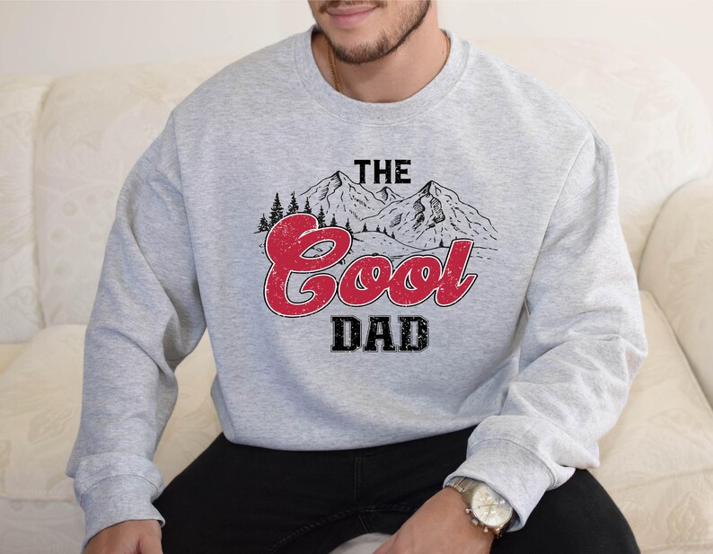 May include: A light gray sweatshirt with the text "THE Cool DAD" in a red and black design. The graphic includes a mountain range and trees. The sweatshirt is long-sleeved and made of a soft material.