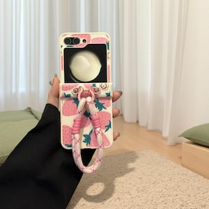 May include: A white and pink phone case featuring a strawberry design. The case includes a strap made of pink and white rope. The phone case is open, showing a mirror.