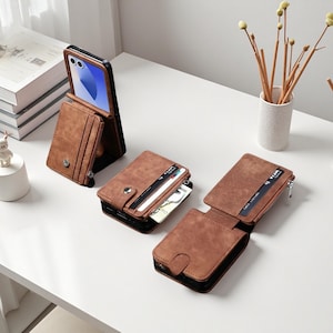 May include: Brown leather wallet with a phone holder, featuring a snap closure and a zippered compartment. The wallet is designed to hold cards and cash. The phone is displayed in a vertical position. The wallet is on a white surface.