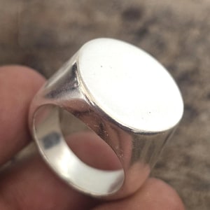 Può includere: Un anello con sigillo d'argento con una faccia ovale lucida. L'anello ha una fascia spessa e una superficie liscia e riflettente. L'anello è un classico gioiello.