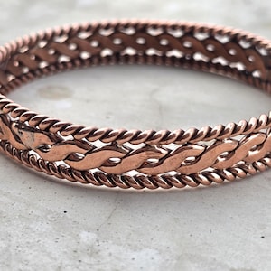 Op de afbeelding: Een koperkleurige armband met een gevlochten ontwerp. De armband heeft een centraal gedeelte van verweven schakels, begrensd door gedraaide touwachtige randen. Het metaal heeft een warme, roodbruine tint en het ontwerp is ingewikkeld en gedetailleerd.