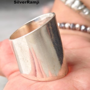 May include: A silver-colored ring with a unique, angled design. The ring has a smooth, polished surface and a cylindrical shape, with one side cut at an angle. The ring is shown on a person's hand.
