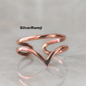 May include: A copper-colored ring with a V-shaped design. The ring has a double-banded structure, with the V-shape at the front. The text "SilverRamji" is printed on the top left of the image. The ring is displayed on a reflective surface.