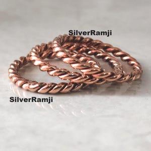May include: Close-up of three copper-colored twisted rope rings. The rings are stacked on top of each other, with the text "SilverRamji" in black on the top and bottom of the image. The rings have a shiny finish.