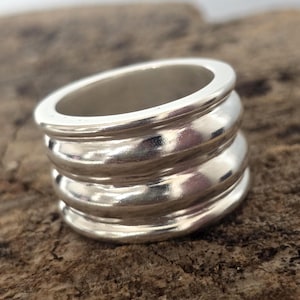 Puede incluir: Un anillo plateado con una banda gruesa con tres crestas redondeadas apiladas. El anillo tiene una superficie pulida y reflectante y se asienta sobre una superficie de madera marrón texturizada. Es una joya.