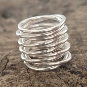 Può includere: Un anello d'argento composto da più fasce avvolte. L'anello ha una superficie liscia e lucida e un aspetto luminoso e riflettente. L'anello è posto su uno sfondo marrone strutturato, evidenziando la sua lucentezza metallica. Un gioiello.