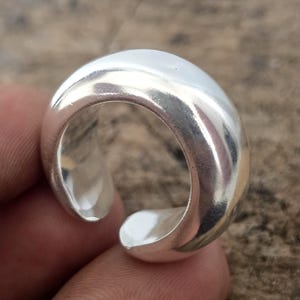 Puede incluir: Un anillo plateado brillante con un diseño redondeado y liso. El anillo tiene una banda gruesa y un acabado pulido, que refleja la luz. Una joya sencilla pero elegante.