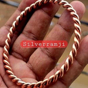 May include: A copper-coloured twisted bangle bracelet. The bracelet is made of intertwined strands, creating a rope-like appearance. The text "Silverramji" is displayed in a red rectangle.