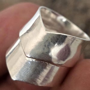 Puede incluir: Primer plano de un anillo plateado con un diseño geométrico. El anillo tiene una apariencia en capas con superficies angulares y pulidas. El metal refleja la luz, creando reflejos y sombras. El anillo es una joya.