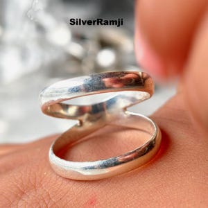 Könnte beinhalten: Nahaufnahme eines Silberrings mit einem einzigartigen Doppelband-Design. Der Ring hat eine polierte, reflektierende Oberfläche, und die Bänder kreuzen sich. Der Ring befindet sich auf der Haut einer Person. Der Text "SilverRamji" befindet sich oben in der Mitte.