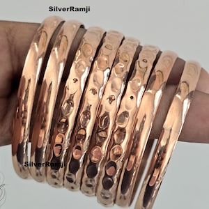 May include: A collection of copper bangles, some smooth and others with a hammered texture, are displayed. The bangles are a warm, reddish-brown color and appear to be made of metal. The text "SilverRamji" is visible in the image.