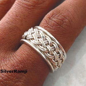Op de afbeelding: Een zilveren ring met een gevlochten ontwerp. De ring is breed en heeft een gedetailleerd patroon van verweven metalen strengen. De ring wordt om een vinger gedragen en toont het ingewikkelde vakmanschap en het heldere, reflecterende oppervlak van het metaal.