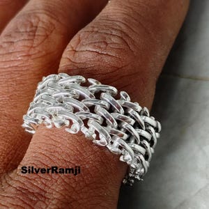 Puede incluir: Un anillo de plata con un diseño de cadena trenzada. El anillo es de metal brillante y tiene una superficie texturizada. El anillo está en un dedo. El texto "SilverRamji" es visible en la imagen.
