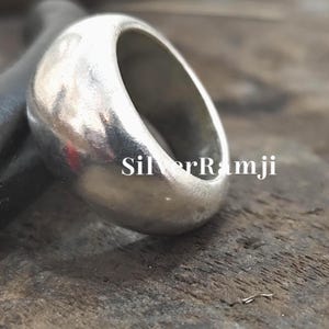 Puede incluir: Primer plano de un anillo de plata con un diseño redondeado y liso. El anillo tiene una banda ancha y un acabado pulido. El texto "SilverRamji" es visible en la superficie. El anillo es probablemente de plata de ley.