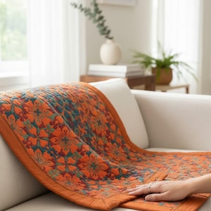 Puede incluir: Un edredón decorativo con un estampado floral naranja y turquesa, sobre un sofá blanco. El edredón presenta un diseño floral repetido y un borde naranja oscuro. La habitación es luminosa, con una ventana y plantas decorativas.