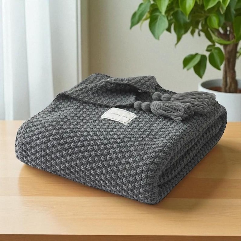 Puede incluir: Una manta gris tejida, doblada, con un patr&oacute;n texturizado y borlas decorativas. La manta est&aacute; cuidadosamente apilada sobre una superficie de madera, con una peque&ntilde;a etiqueta adjunta. Una planta de interior es visible en el fondo.