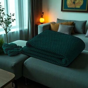 Puede incluir: Manta verde oscuro plegada con un patr&oacute;n texturizado, sobre un sof&aacute; seccional gris. Se ve una borla verde a juego. La manta es un elemento de decoraci&oacute;n del hogar.