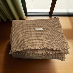 May include: A folded, tan-colored blanket with a textured, grid-like pattern. The blanket has fringed edges and a small, rectangular tag in the center. The blanket is made of a soft material and is folded neatly.