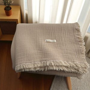 May include: A beige, textured blanket with fringed edges is draped over a wooden chair. The blanket has a label in the center. The blanket is made of a soft material and is suitable for home decor.
