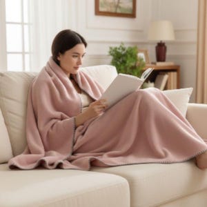 Op de afbeelding: Een vrouw zit op een crèmekleurige bank, gehuld in een zachte, stoffige roze deken met grijze rand. Ze leest een boek, wat een gezellige en ontspannen sfeer creëert. De deken lijkt van een zacht materiaal te zijn.